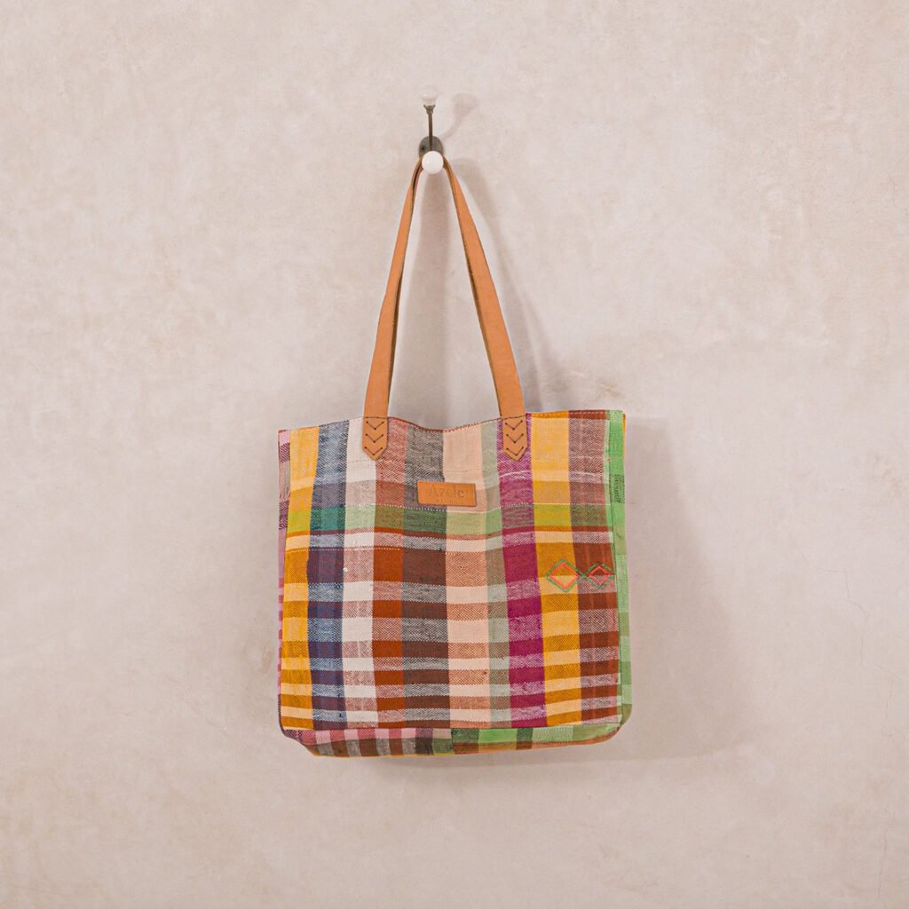 A colorful, plaid-patterned tote bag with brown leather handles hangs on a wall hook.