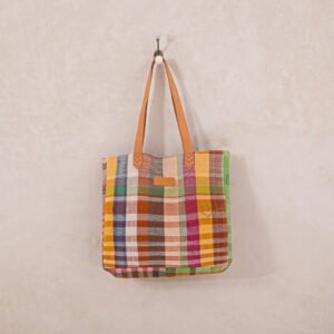 A colorful, plaid-patterned tote bag with brown leather handles hangs on a wall hook.
