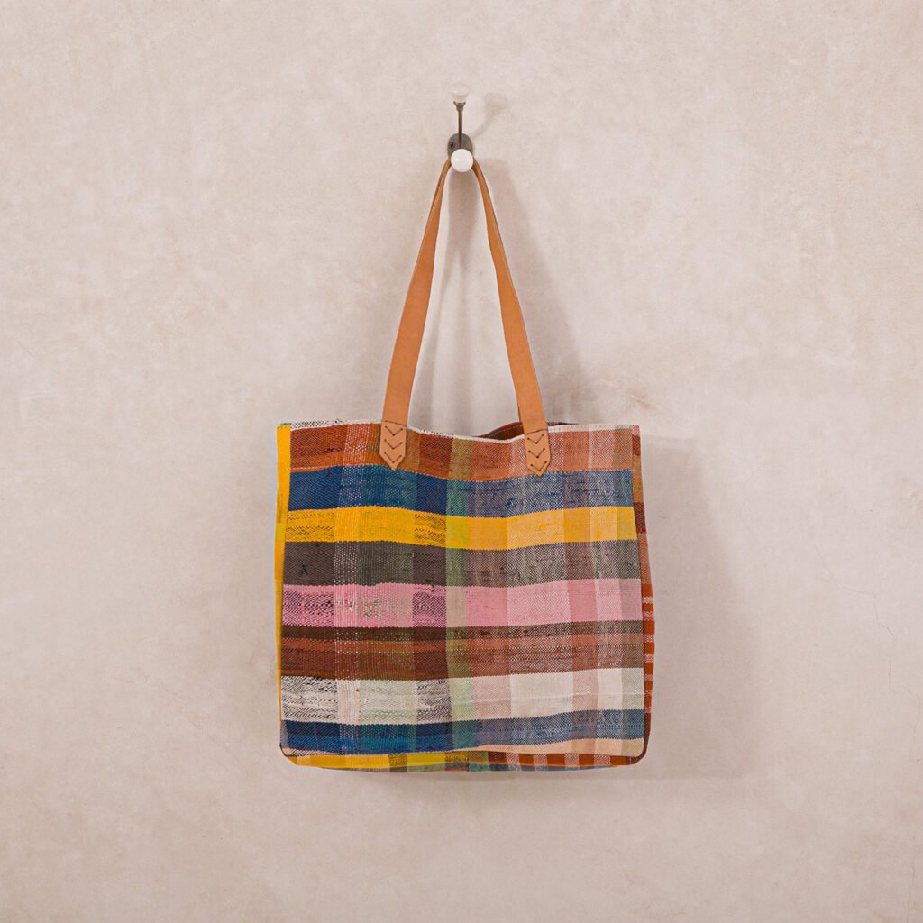 A multicolored checkered tote bag with leather handles hangs on a wall hook.