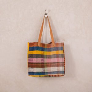 A multicolored checkered tote bag with leather handles hangs on a wall hook.