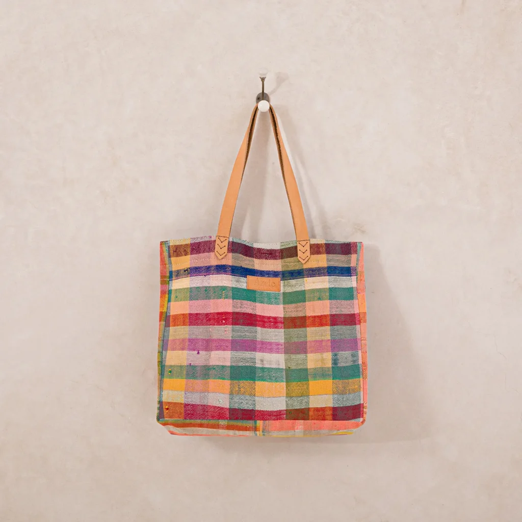 A multicolored, plaid-patterned tote bag with brown leather handles hangs on a wall hook.