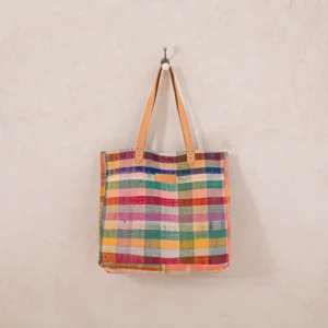 A multicolored, plaid-patterned tote bag with brown leather handles hangs on a wall hook.