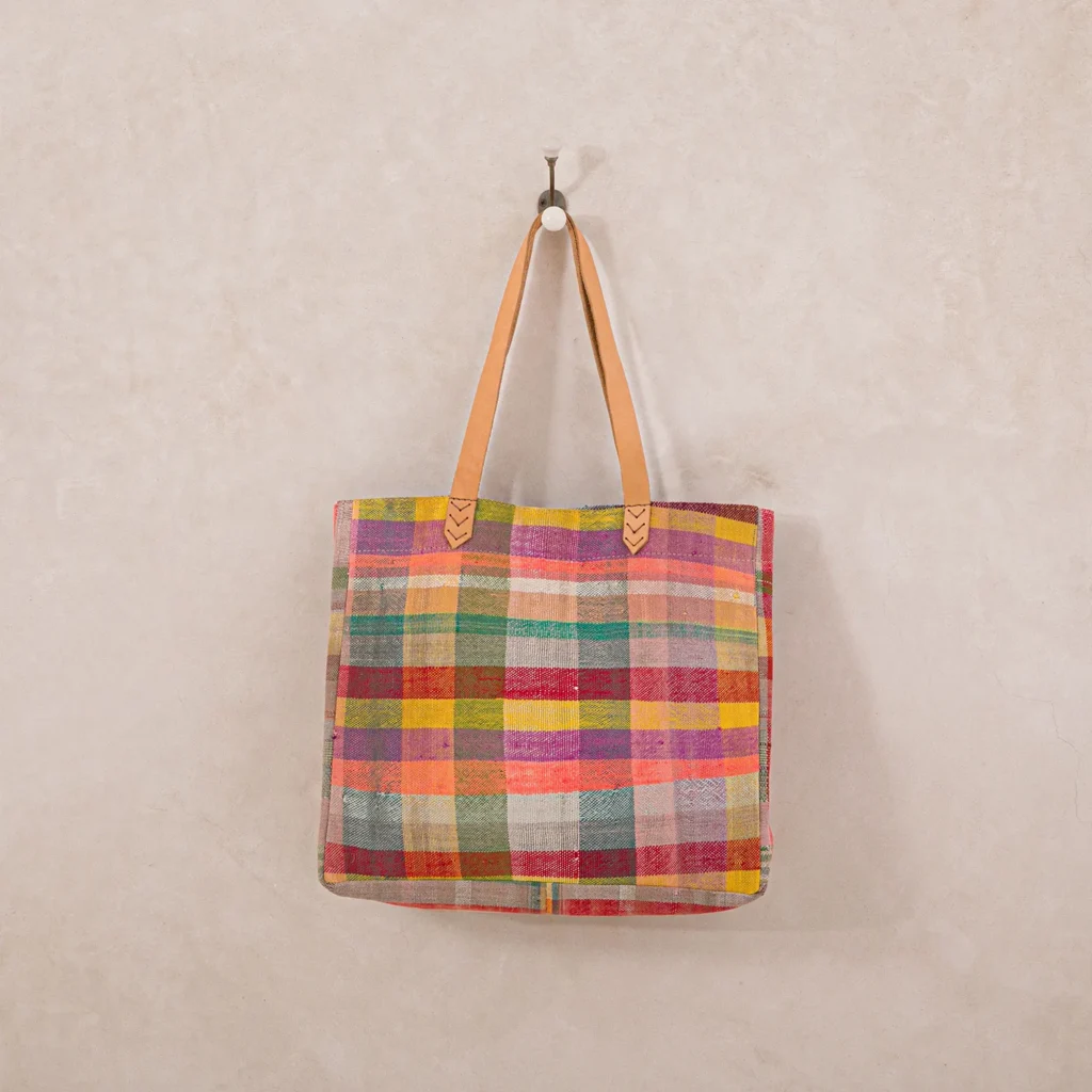 A colorful checkered tote bag with tan leather straps hangs on a single wall hook against a light beige background.