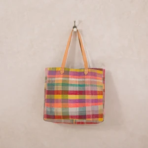 A colorful checkered tote bag with tan leather straps hangs on a single wall hook against a light beige background.