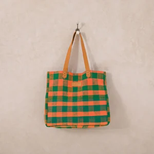 An upcycled shopper bag in green and orange checkered patterns, complete with tan leather handles, hangs elegantly on a wall hook.