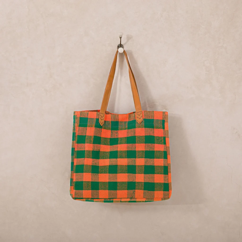 An upcycled shopper bag, with plaid patterns in green and orange checks, hangs on a wall hook by its brown leather straps.