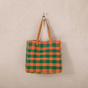 An upcycled shopper bag, with plaid patterns in green and orange checks, hangs on a wall hook by its brown leather straps.