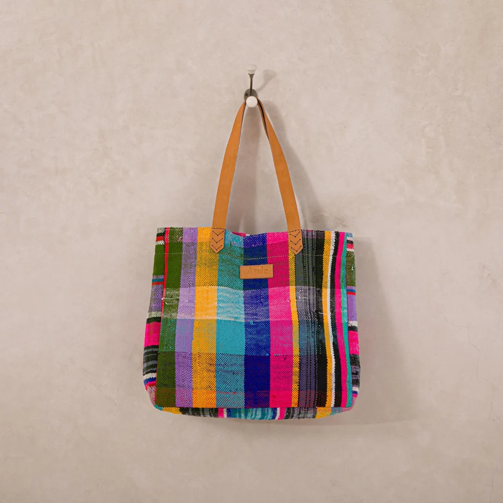Multicolored plaid tote bag with tan leather handles hanging on a wall hook.