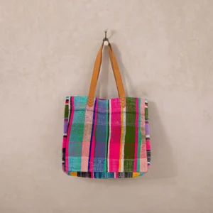 A colorful, checkered fabric tote bag with brown leather handles hangs on a wall hook against a plain beige background.
