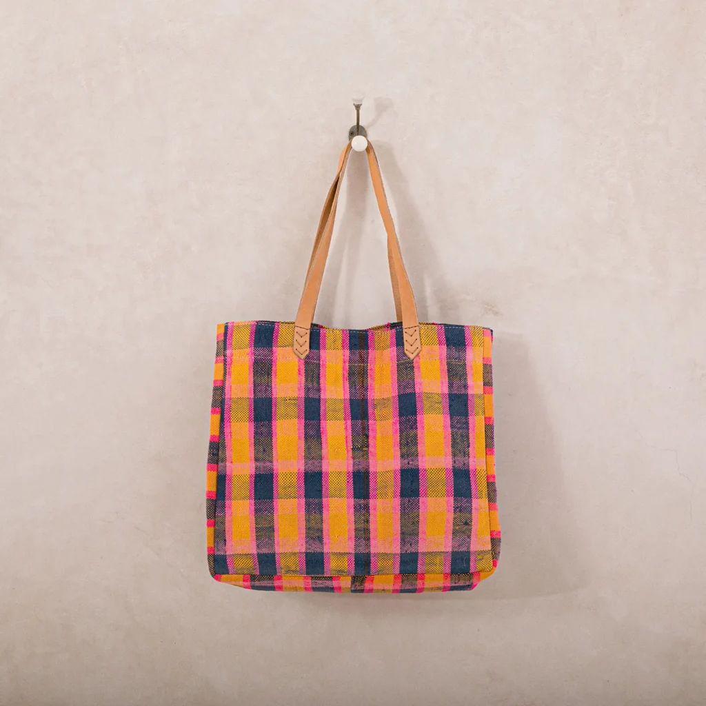 A tote bag with multicolored plaid pattern, mainly pink, yellow, and blue, hangs from a wall hook.