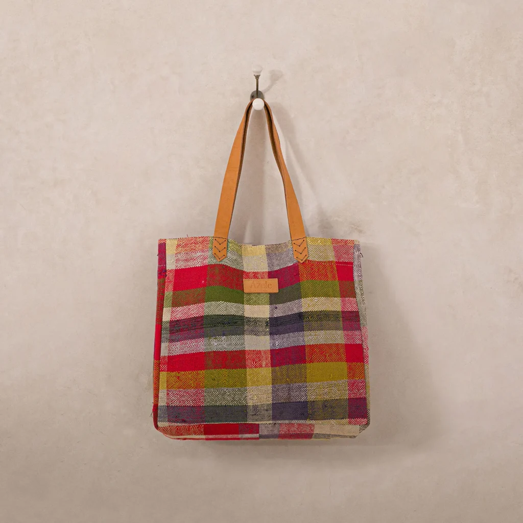 Plaid fabric tote bag with brown leather handles hanging on a hook against a plain wall.