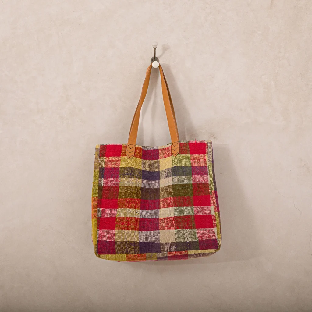 A colorful plaid tote bag with red, green, yellow, and purple squares, hangs by its brown leather straps on a beige wall.