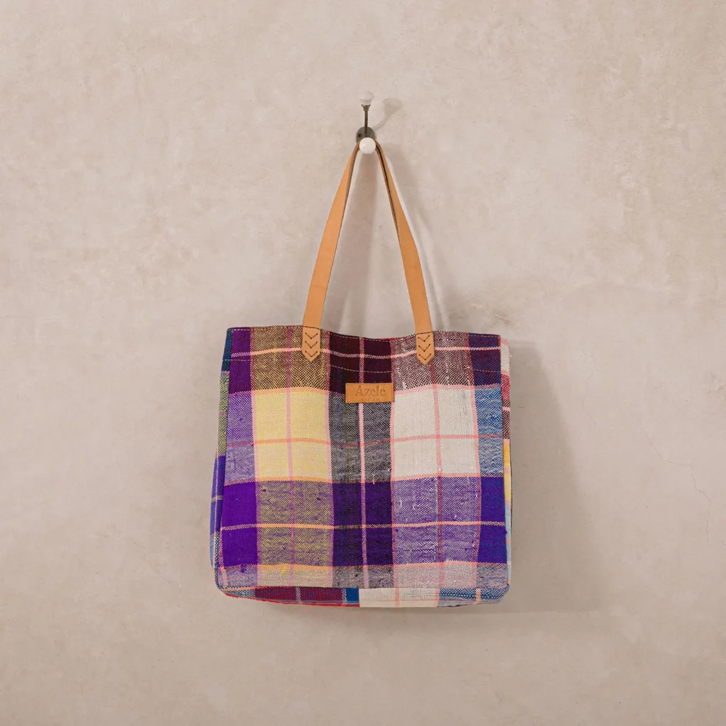 A multicolored plaid tote bag with tan leather handles hangs on a hook against a beige wall.