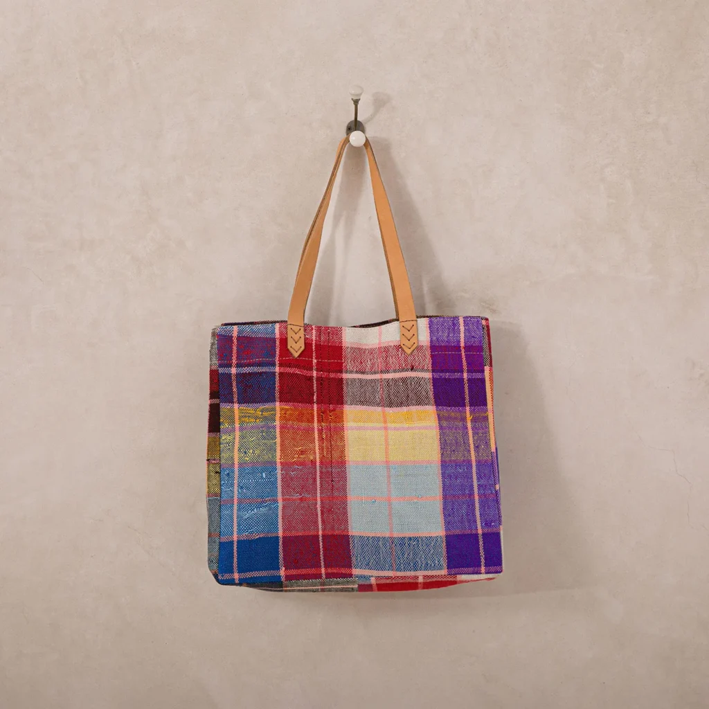 A colorful, plaid-patterned tote bag with leather handles hangs on a simple wall hook.