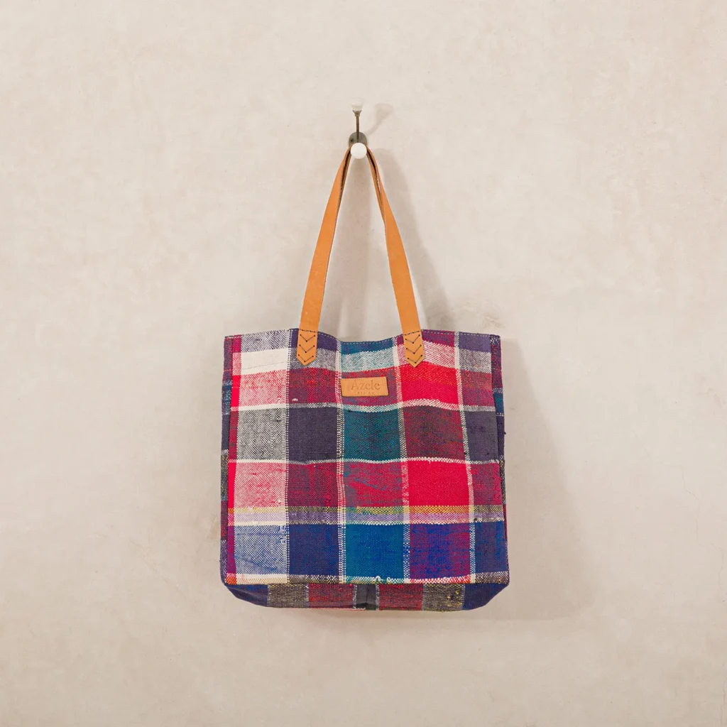 A plaid fabric tote bag with brown leather handles hangs on a metal hook against a beige textured wall.