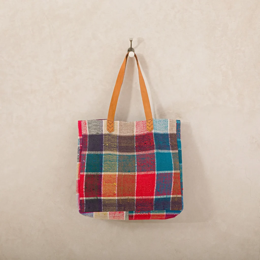 A multi-colored patchwork tote bag with brown leather handles hangs on a hook against a plain, textured wall.