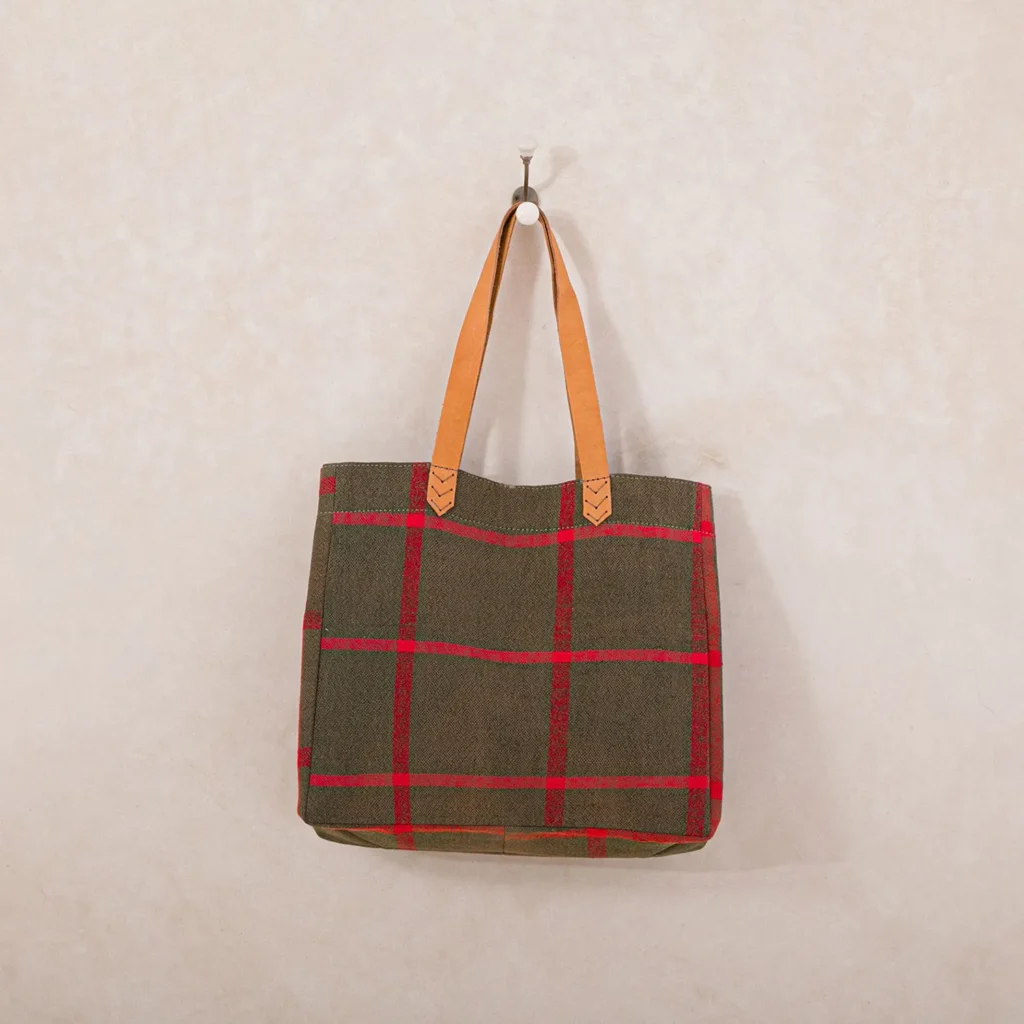 A tote bag with red plaid pattern and brown leather handles hangs on a wall hook against a textured beige background.