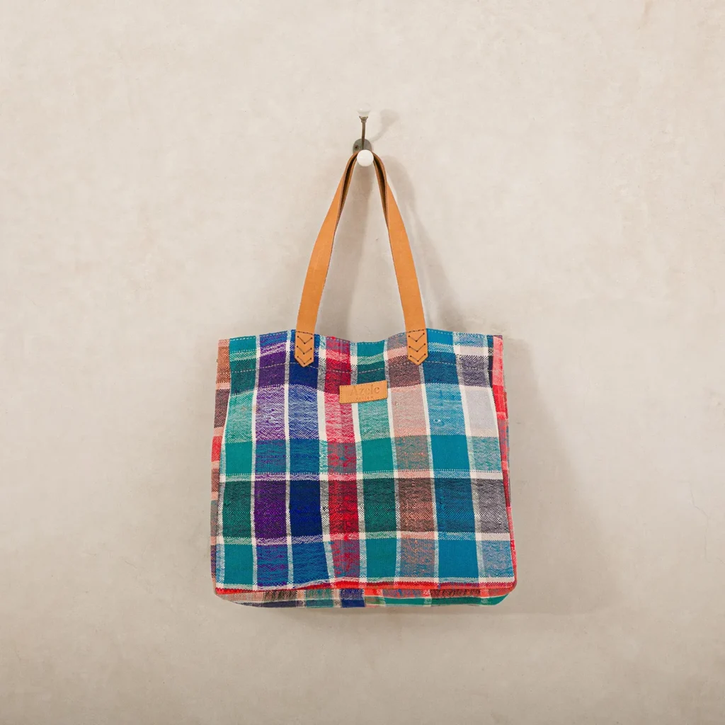 A multicolored plaid tote bag with brown leather handles hangs on a wall hook.