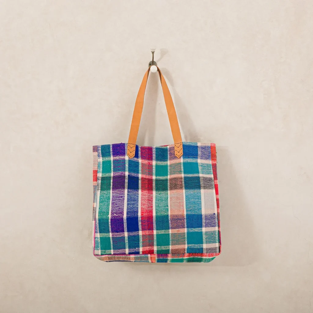 A multicolored plaid tote bag with brown leather handles hangs on a wall hook against a beige background.