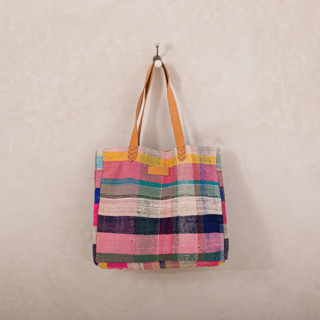 A multicolored, checkered fabric tote bag with tan leather handles hangs on a single hook against a plain wall.