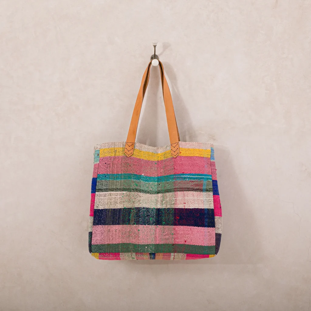 A colorful woven tote bag with leather handles hangs on a wall hook. The design features horizontal stripes in pink, blue, green, and yellow.