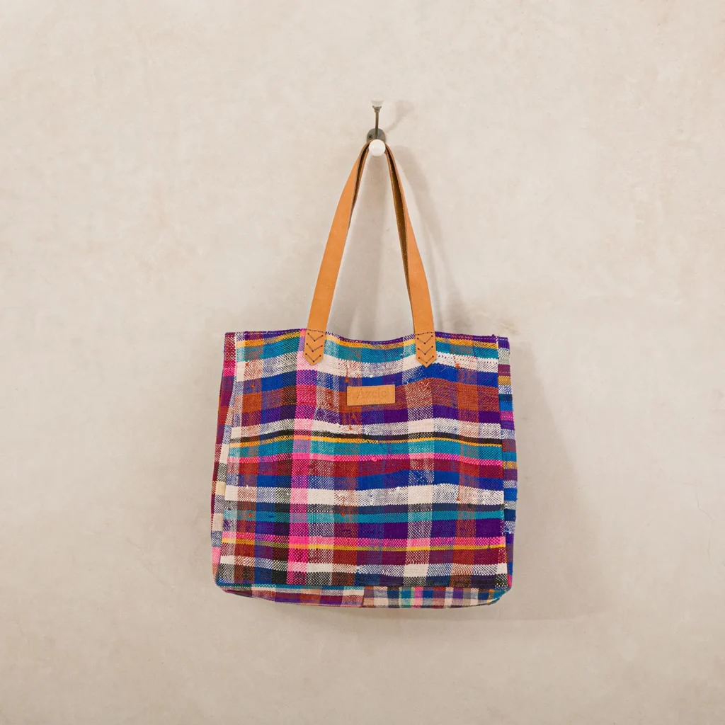 A colorful woven tote bag with a plaid pattern and tan leather handles hangs on a wall hook.