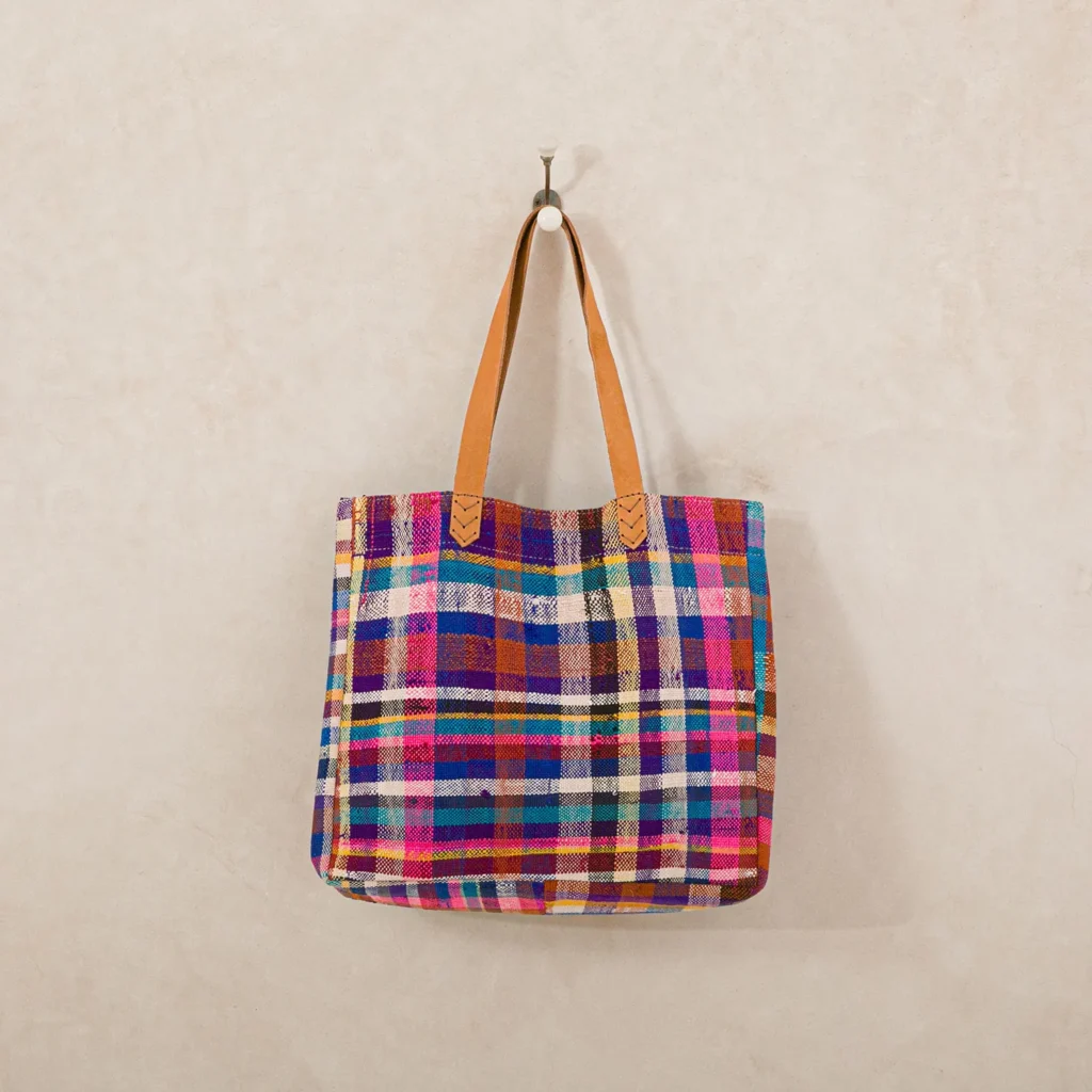 A multicolored plaid tote bag with tan leather straps hangs on a wall hook.