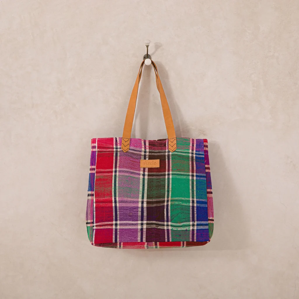 Plaid tote bag with red, green, purple, and blue patterns, hanging on a wall hook. Brown leather handles are attached to the bag.