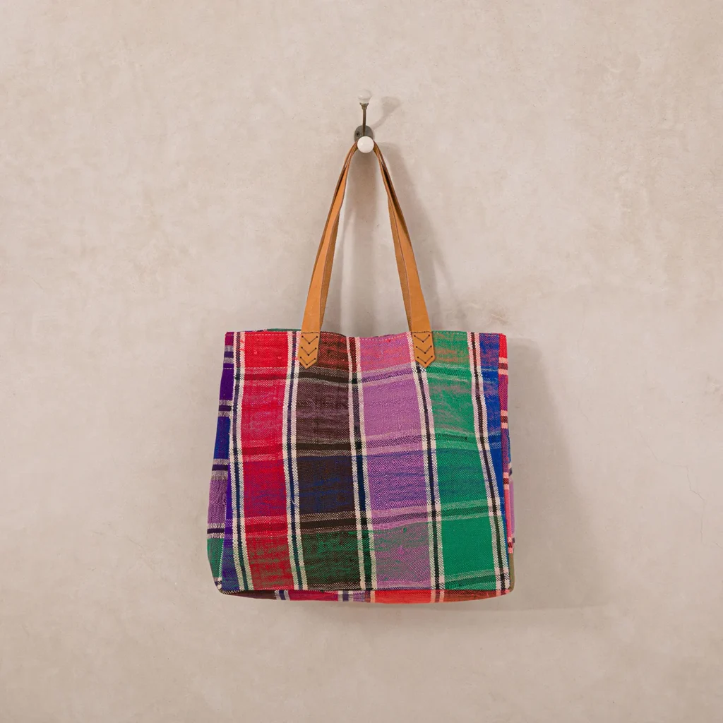A multicolored plaid tote bag with brown leather handles hangs on a wall hook.