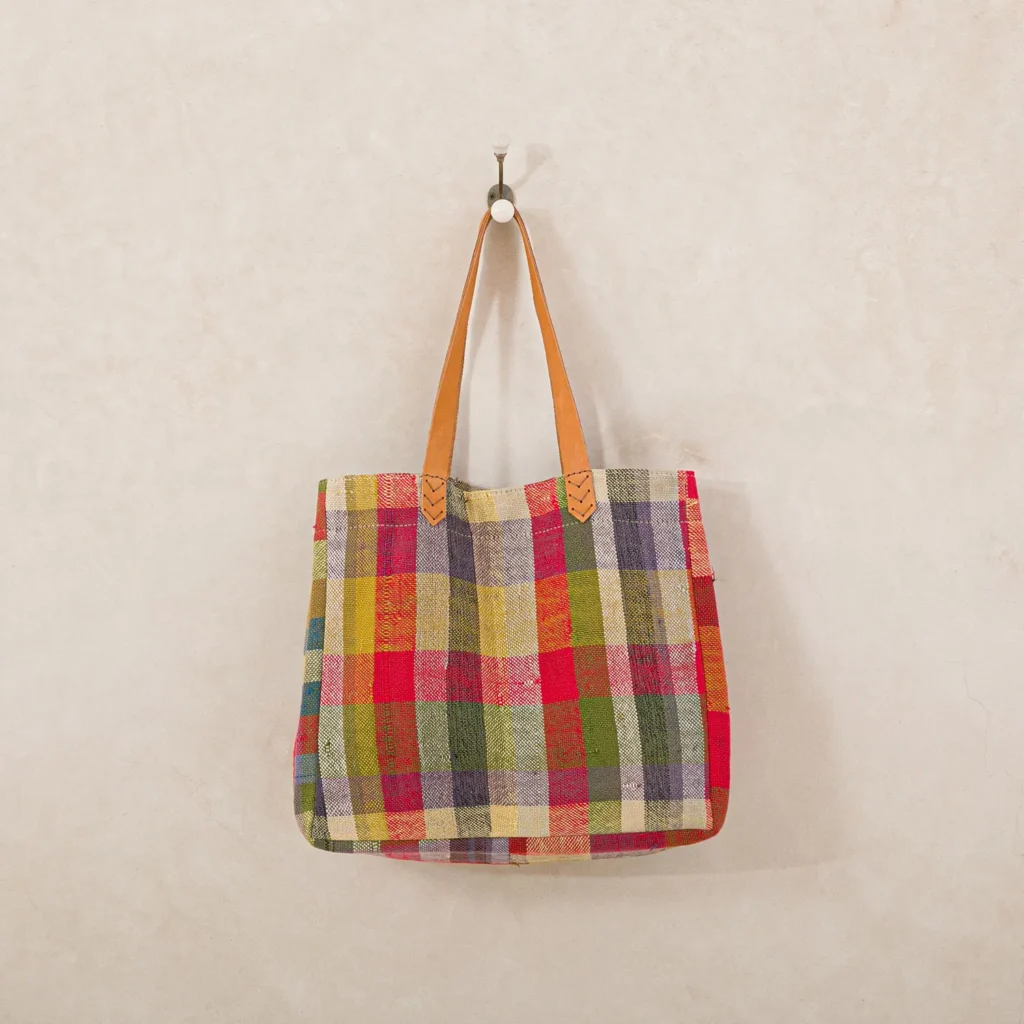 A vibrant, multicolored plaid tote bag with brown leather straps hangs elegantly on a single wall hook against a plain background, adding a touch of stylish geometry to the room.