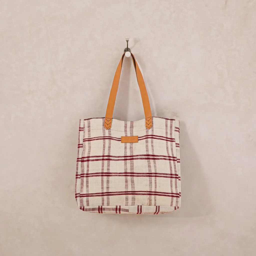 A plaid tote bag with red and beige checks hangs on a wall hook, showcasing its brown leather straps and minimalist design. This stylish accessory subtly complements any modern decor without overpowering it.