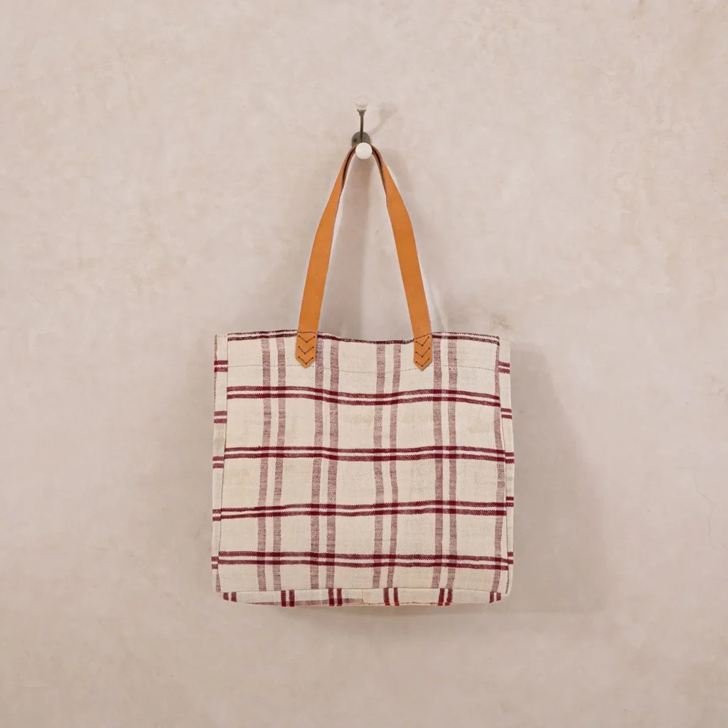 A stylish plaid tote bag with a red and white checkered pattern and brown leather handles hangs elegantly on a wall hook, perfect for adding charm to any space.