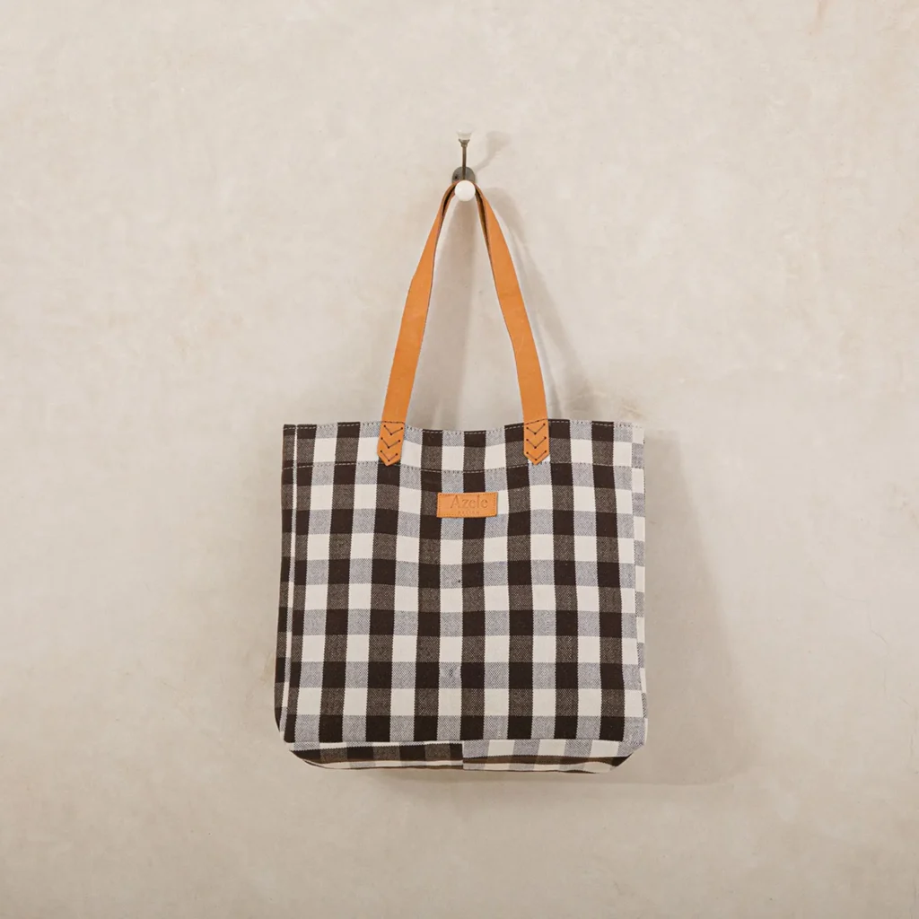 A black and white checkered tote bag with brown leather handles hangs on a wall hook against a light beige background.