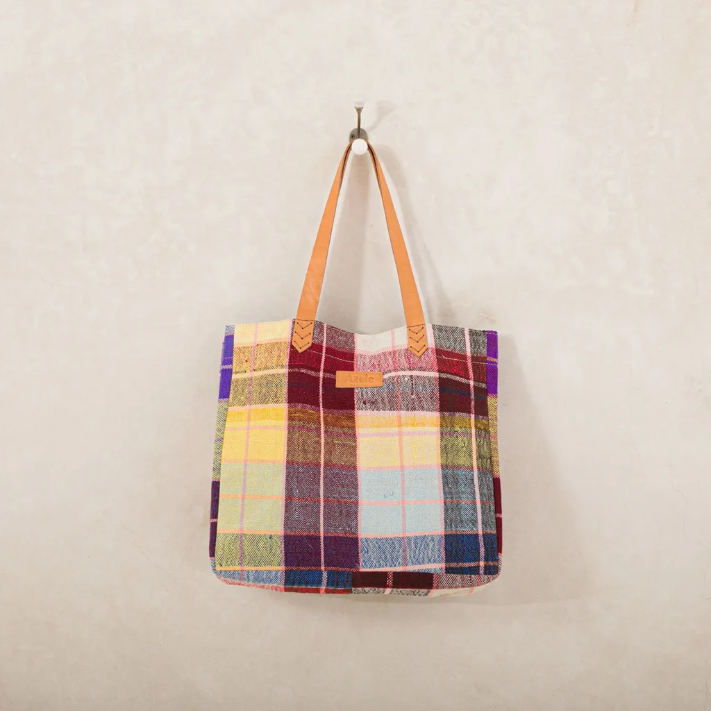 A multicolored plaid tote bag with brown leather handles hangs on a hook against a light textured wall.