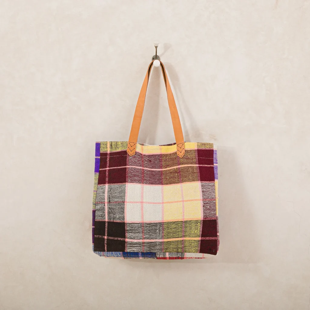 A plaid tote bag with brown leather handles, hanging on a metal hook against a beige wall.