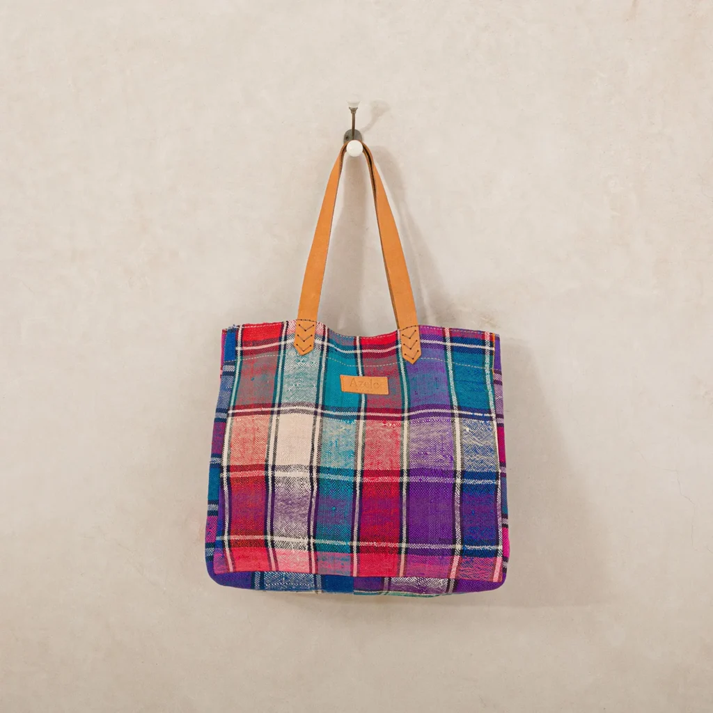 A colorful plaid tote bag with leather straps hangs on a single wall hook against a neutral wall.