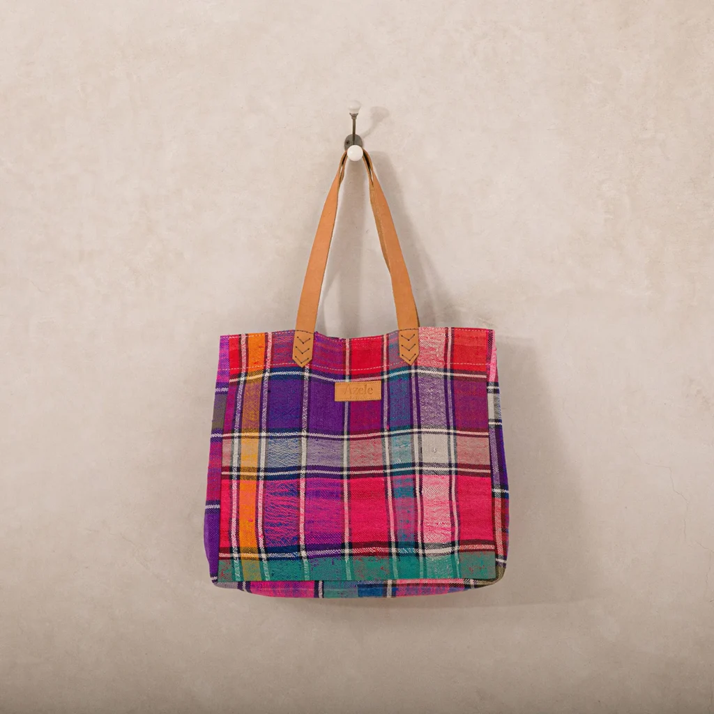 A colorful, plaid-patterned tote bag with brown handles hangs on a wall hook against a beige background.