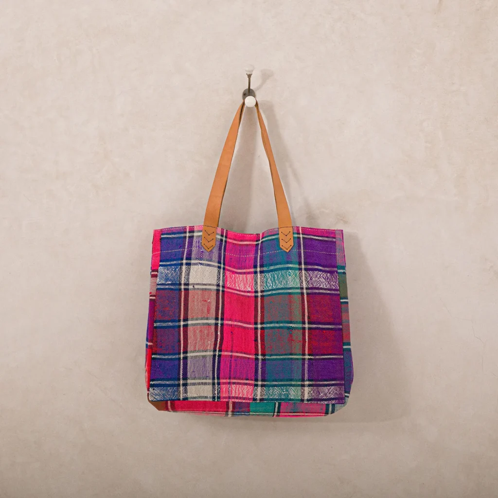 A multicolored plaid tote bag with tan leather handles hangs on a wall hook against a beige background.