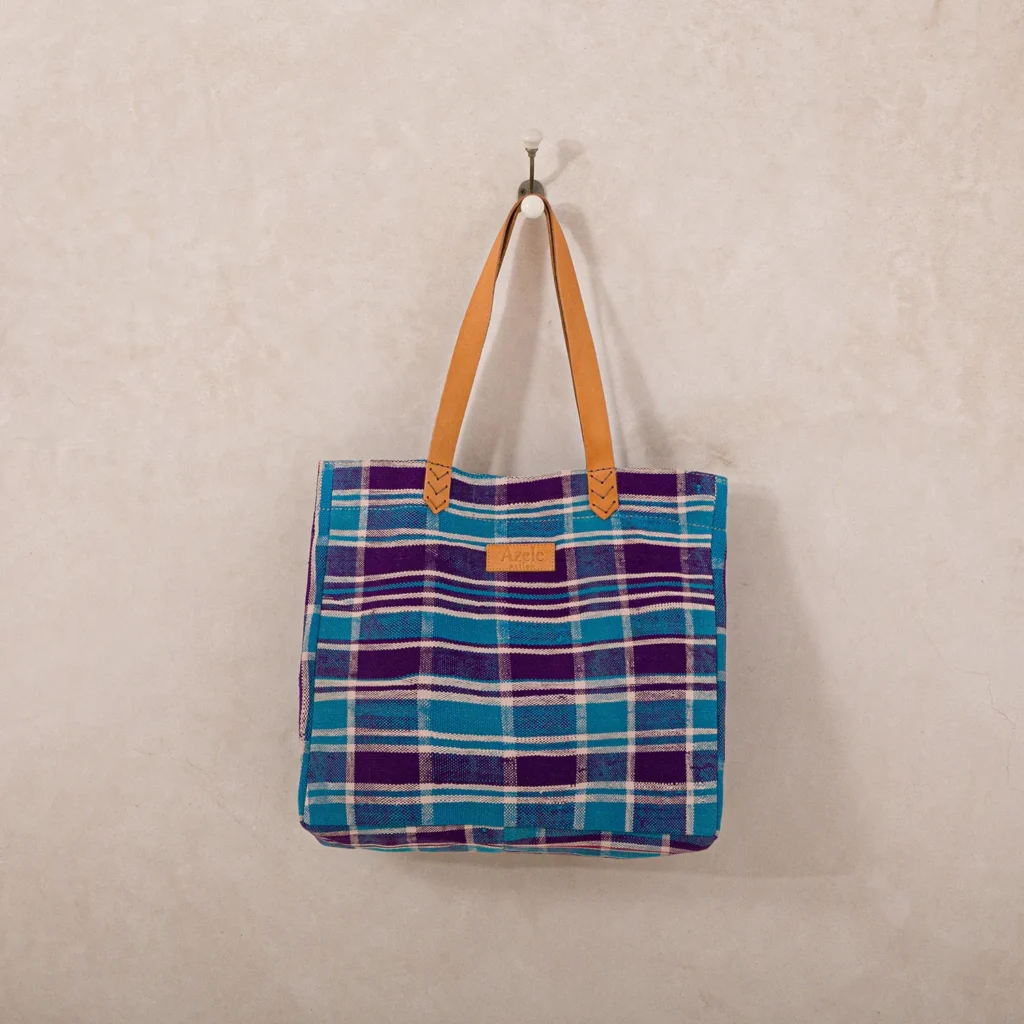 Plaid-patterned tote bag with blue, purple, and green stripes, featuring tan leather straps, hanging on a hook against a beige wall.
