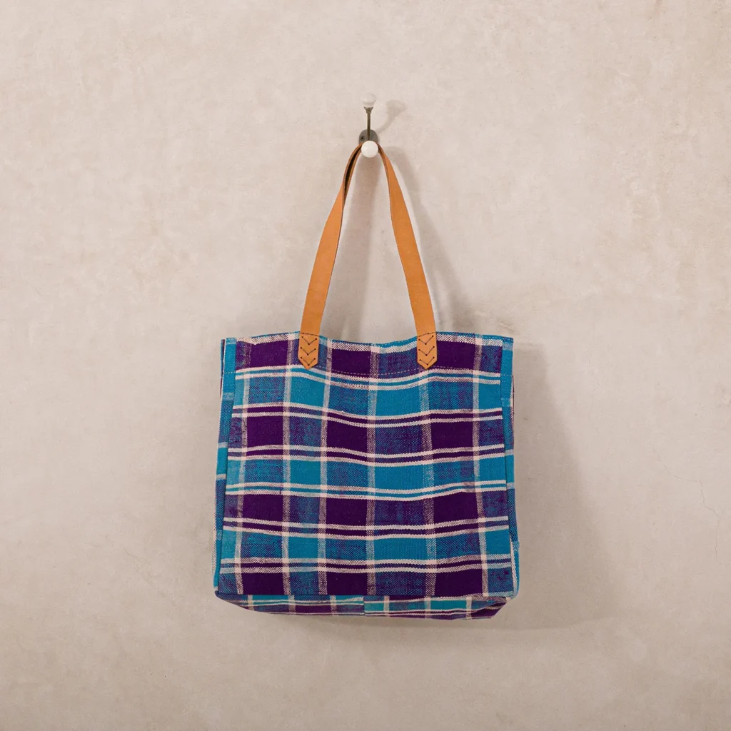 A blue and purple plaid tote bag with brown leather handles hangs on a wall hook.