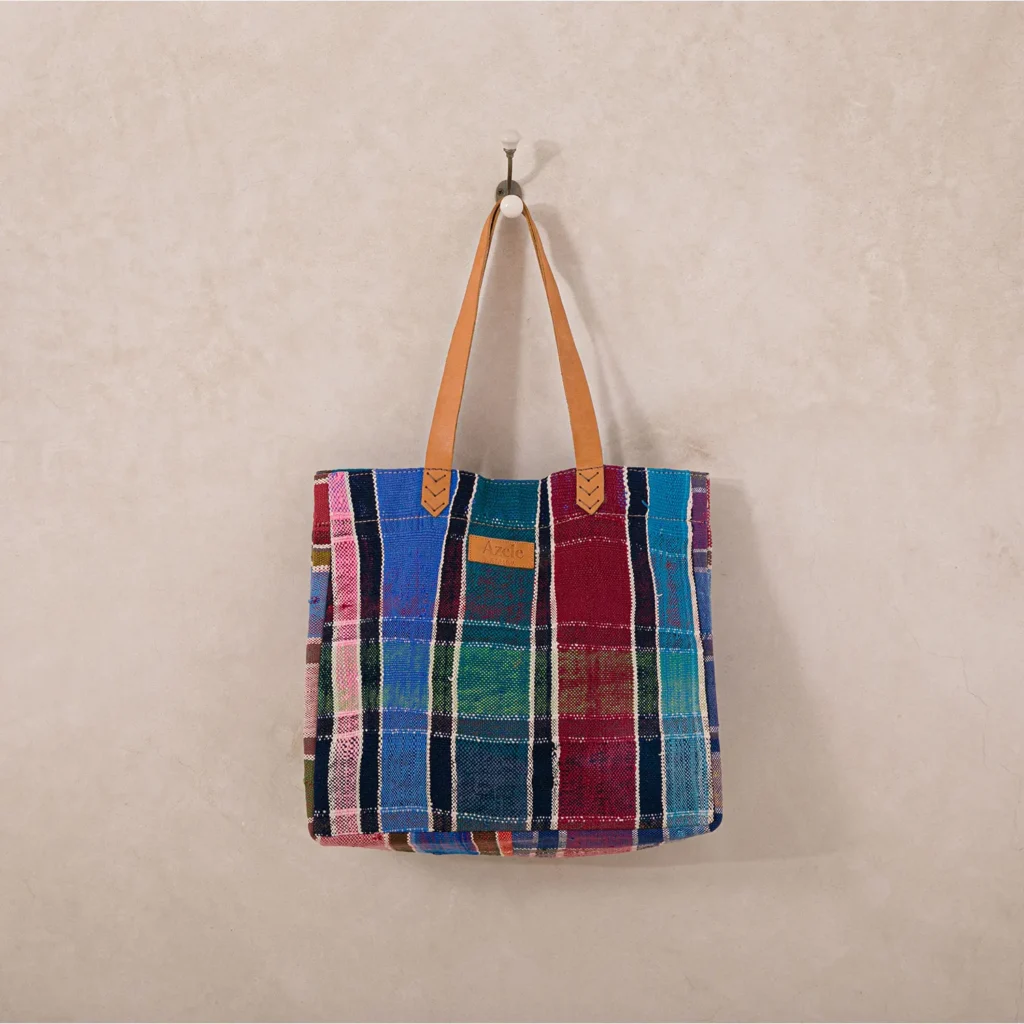 A colorful plaid tote bag with red, blue, green, and beige patterns hangs on a white wall using a leather strap.