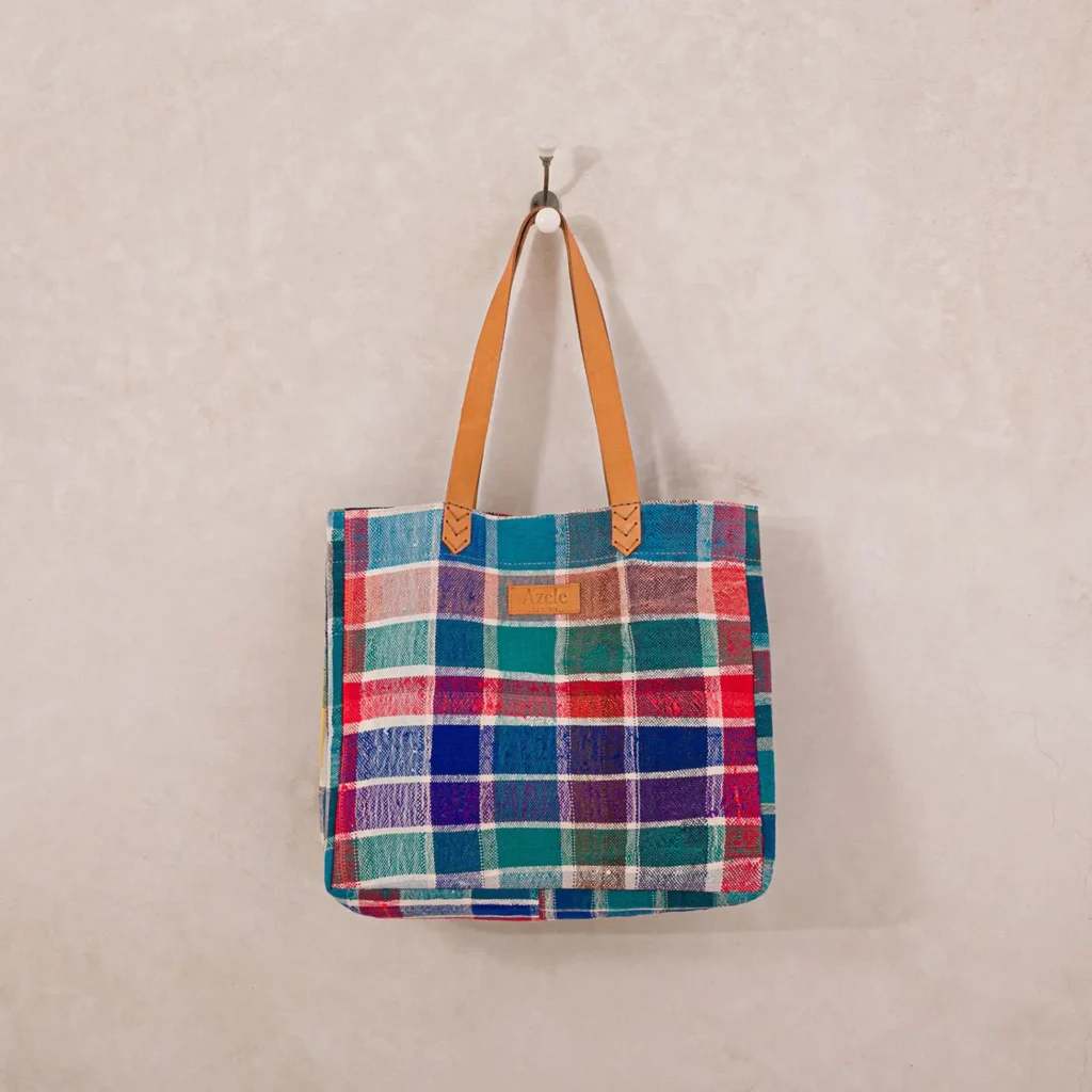 A multicolored plaid tote bag with brown leather handles hangs on a wall hook.