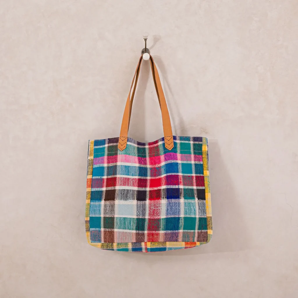 A colorful plaid tote bag with leather handles hangs on a wall hook.