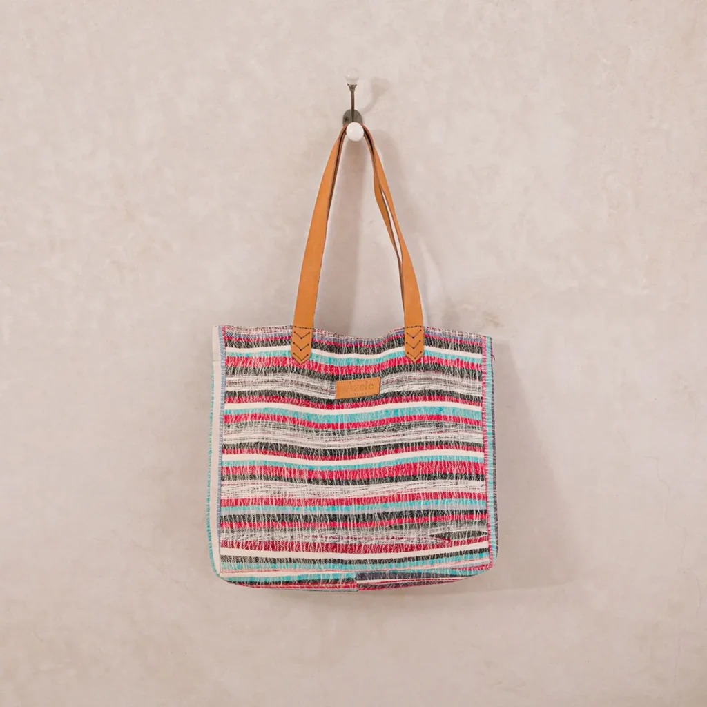 A multicolored striped tote bag with tan leather handles hangs on a simple wall hook against a beige textured background.