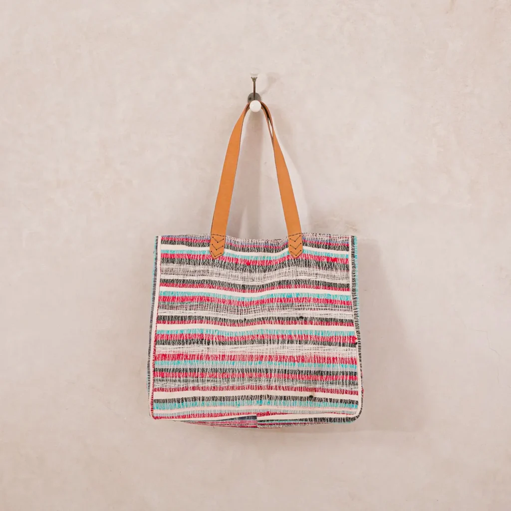 A multicolored striped tote bag with tan leather handles hangs on a white hook against a plain wall.
