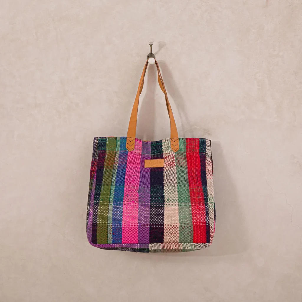 A vibrant woven tote bag featuring tan leather handles elegantly hangs on a wall hook, adding a burst of color to the room.