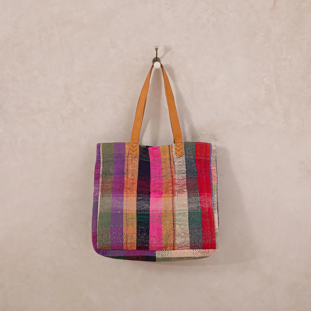 A woven tote bag with a multicolored striped design and tan leather handles hangs on a single hook against a plain beige wall, showcasing its vibrant patterns.