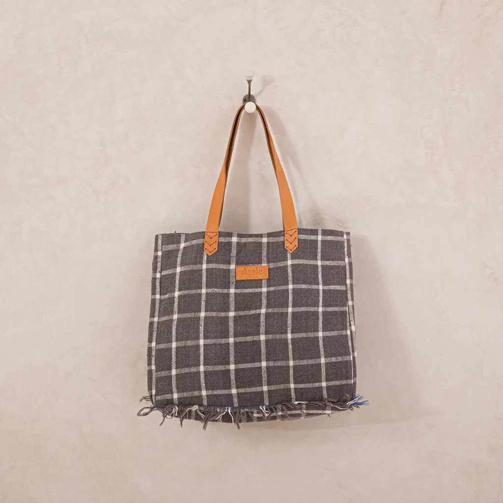 A checkered tote bag with brown leather handles and a leather patch hangs on a wall hook.