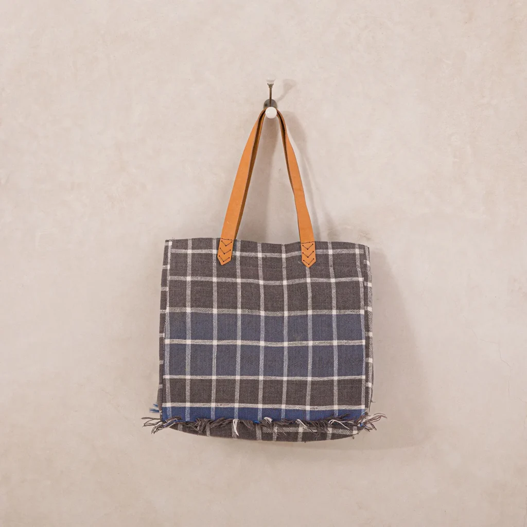 A plaid tote bag with a blue and gray checkered pattern, featuring brown leather handles and frayed edges, hangs on a wall hook.