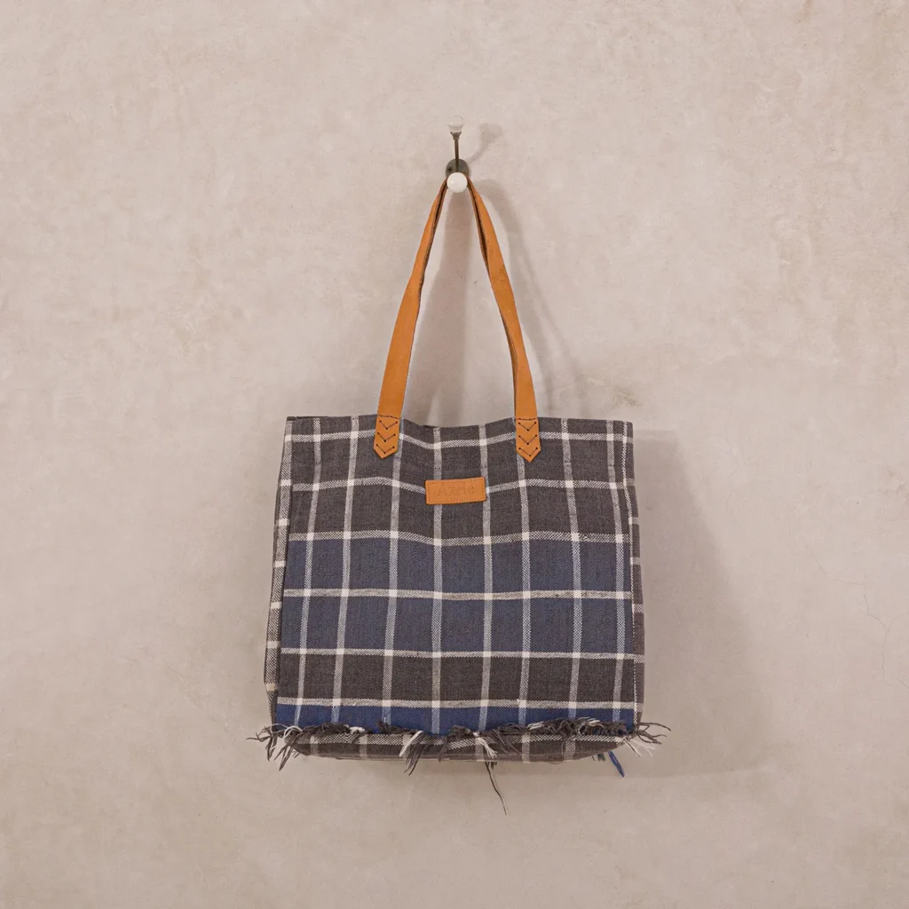 A chic plaid tote bag with frayed edges and brown leather handles gracefully hangs on a wall hook, adding a touch of rustic charm.